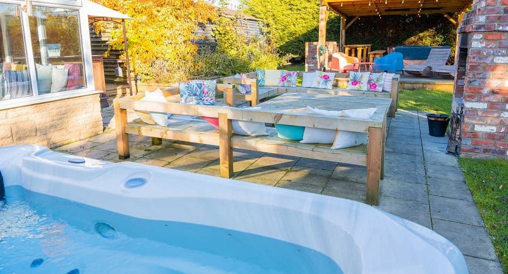 The expansive outdoor social space at Tranquil Escape, a detached Newcastle hen do house, featuring a modern hot tub, rustic social seating for 22, and a garden pergola with a fire pit.